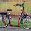 Rode elektrische loopfiets Sitgo Ultimate met lage instap, zadel en accu, geparkeerd buiten op een stoep langs het water.