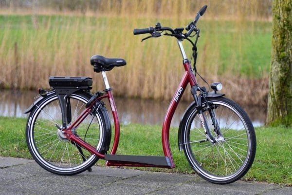Rode elektrische loopfiets Sitgo Ultimate met lage instap, zadel en accu, geparkeerd buiten op een stoep langs het water.